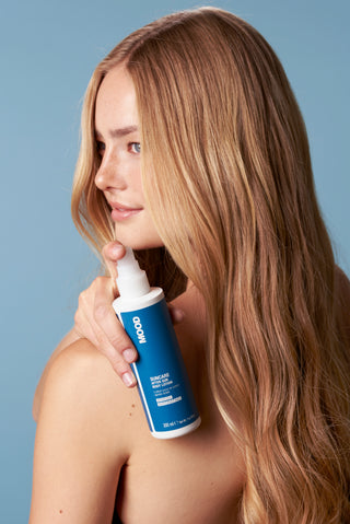 A person holding a bottle of MOOD Suncare After Sun Body Lotion 200ml. The bottle is blue and white, and the person is standing against a blue background.