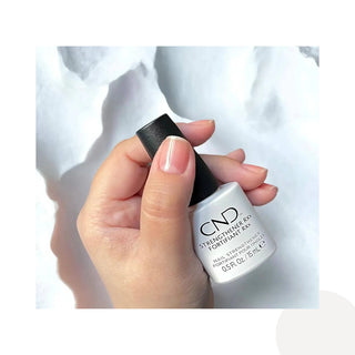 A hand holding a bottle of CND Strengthener RXX Fortifiant RXX nail strengthener, showing a clear bottle with a black cap and a light white liquid inside, set against a white marble background.
