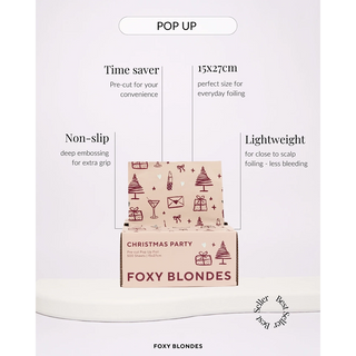 A boxed hair coloring foil labeled Christmas Party and Foxy Blondes, shown on a white pedestal with marketing text around it. The box is pale pink with dark red holiday illustrations and the brand/product name visible on the front.