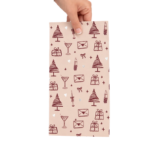 A hand holding a rectangular piece of foil  with a light blush pink background and a repeating dark burgundy holiday pattern of Christmas trees, envelopes, gifts, cocktail glasses, and small stars. 