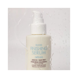 A bottle of Vegan Gloss Finishing Serum with a white pump dispenser, set against a textured white background with water droplets.