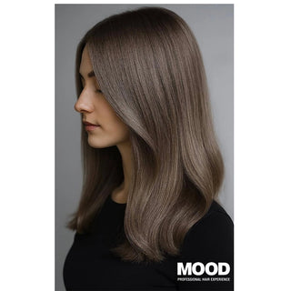 A woman shown in side profile with shoulder-length, straight-to-wavy brown hair featuring subtle ash gray highlights. She is wearing a black top against a plain gray studio background.