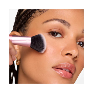 A close-up of a woman's face with makeup being applied using a pink-handled makeup brush on her cheek.