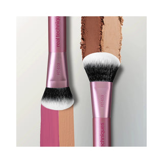 Two Real Techniques makeup brushes in metallic pink and black, shown beside a vertical smear of pressed powder in warm beige and brown shades on a light gray background.