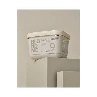 A rectangular cosmetic tub labeled 'BLONDME' and 'PREMIUM LIGHTENER 9+' sits on a geometric pedestal against a warm beige studio background.